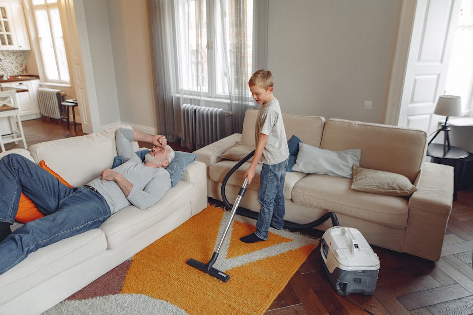 Interior scene of a living room with a wooden floor and area rug, where a yellow vacuum cleaner with a black hose is placed on the rug near a black metal and wooden console table. The table holds a wh