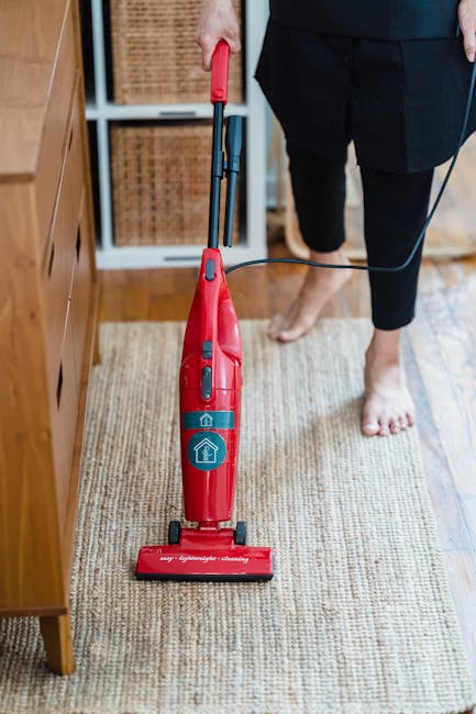 A person standing barefoot on a beige, textured rug in a living room with wooden flooring visible at the edges. The person is operating a red vacuum cleaner with a slim body, mounted on small black wh